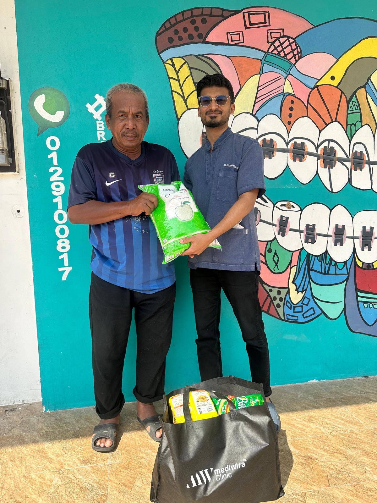 A Mediwira dentist handing a bag of rice and groceries to a resident outside an i-SiHAT Dental clinic
