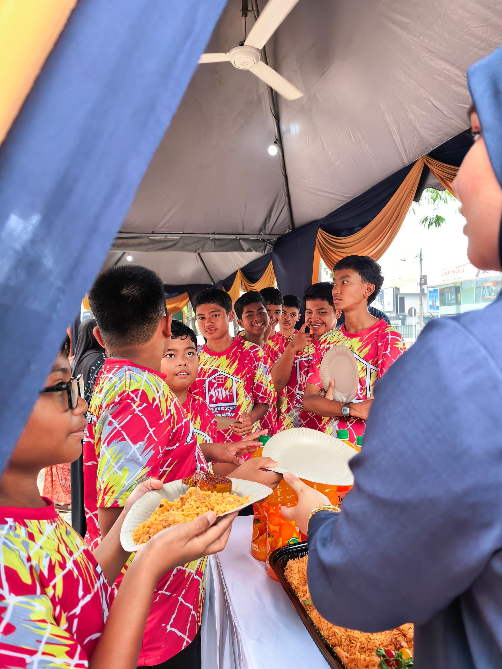 Children queuing for lunch with paper plates and bottles of orange squash under the tent